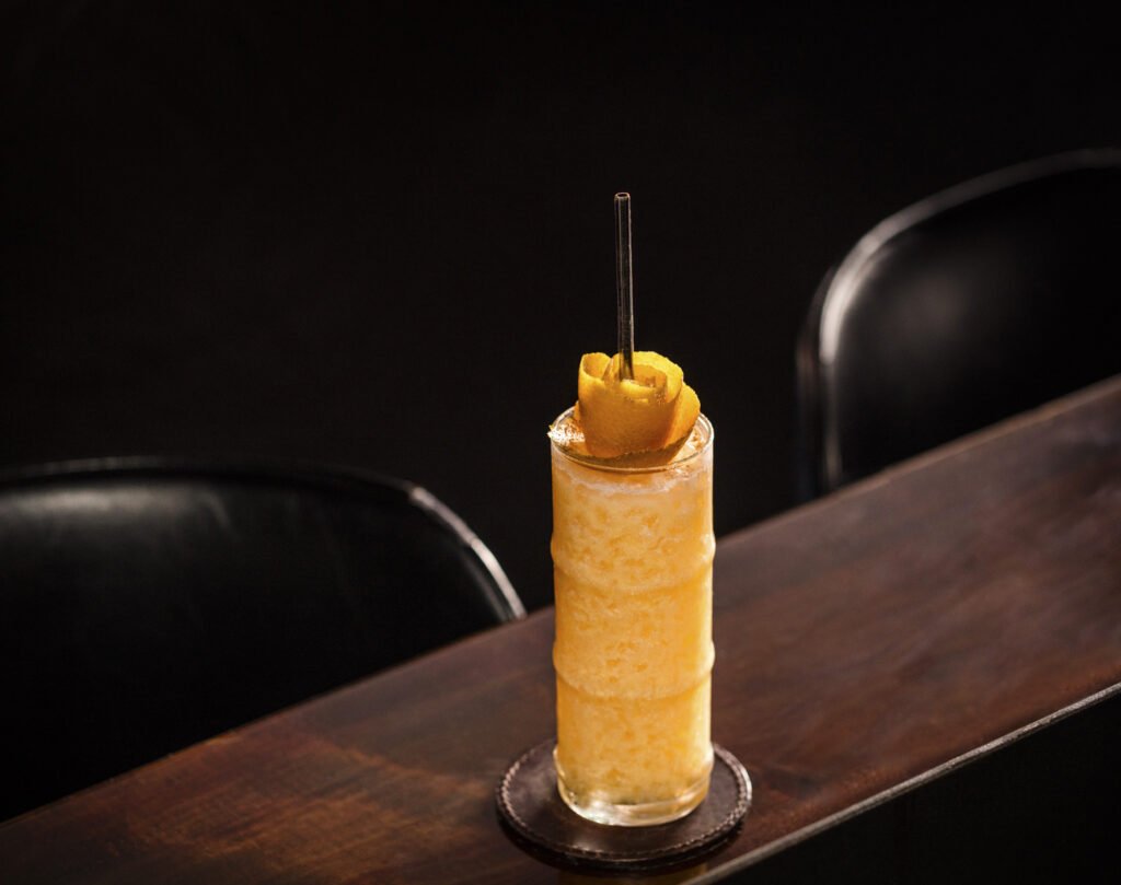 a photo of the Glitter & Marigold mocktail from The Roosevelt Room. A tall bamboo tiki glass to top a table filled with an orange, creamy mocktail, topped with an orange peel rosette and straw. 