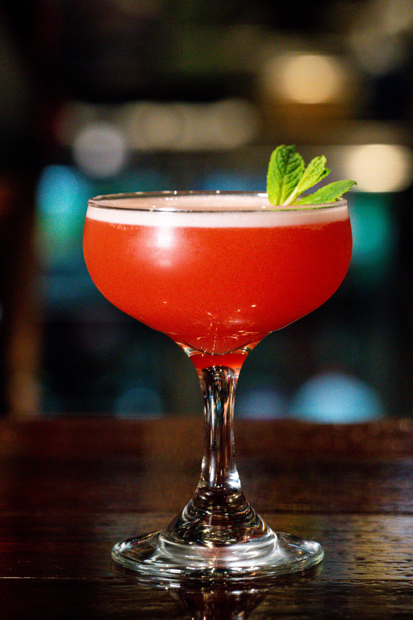 a photo of a Sicilian Punch from Juniper. A red mocktail in a coupe glass on a table garnished with mint leaves